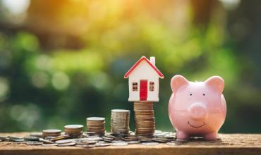 Coins stacked up with a house on top of them and a piggy bank to the right