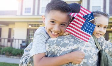 A child holding the US flag being held by their parent in military uniform 