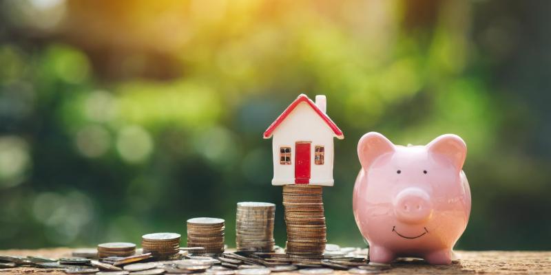 Coins stacked up with a house on top of them and a piggy bank to the right
